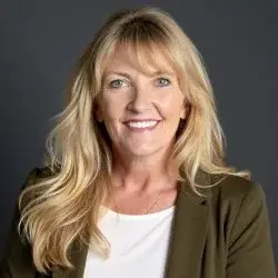 A woman with long blonde hair smiles at the camera. She is wearing a white top and an olive green blazer, standing in front of a plain dark background.